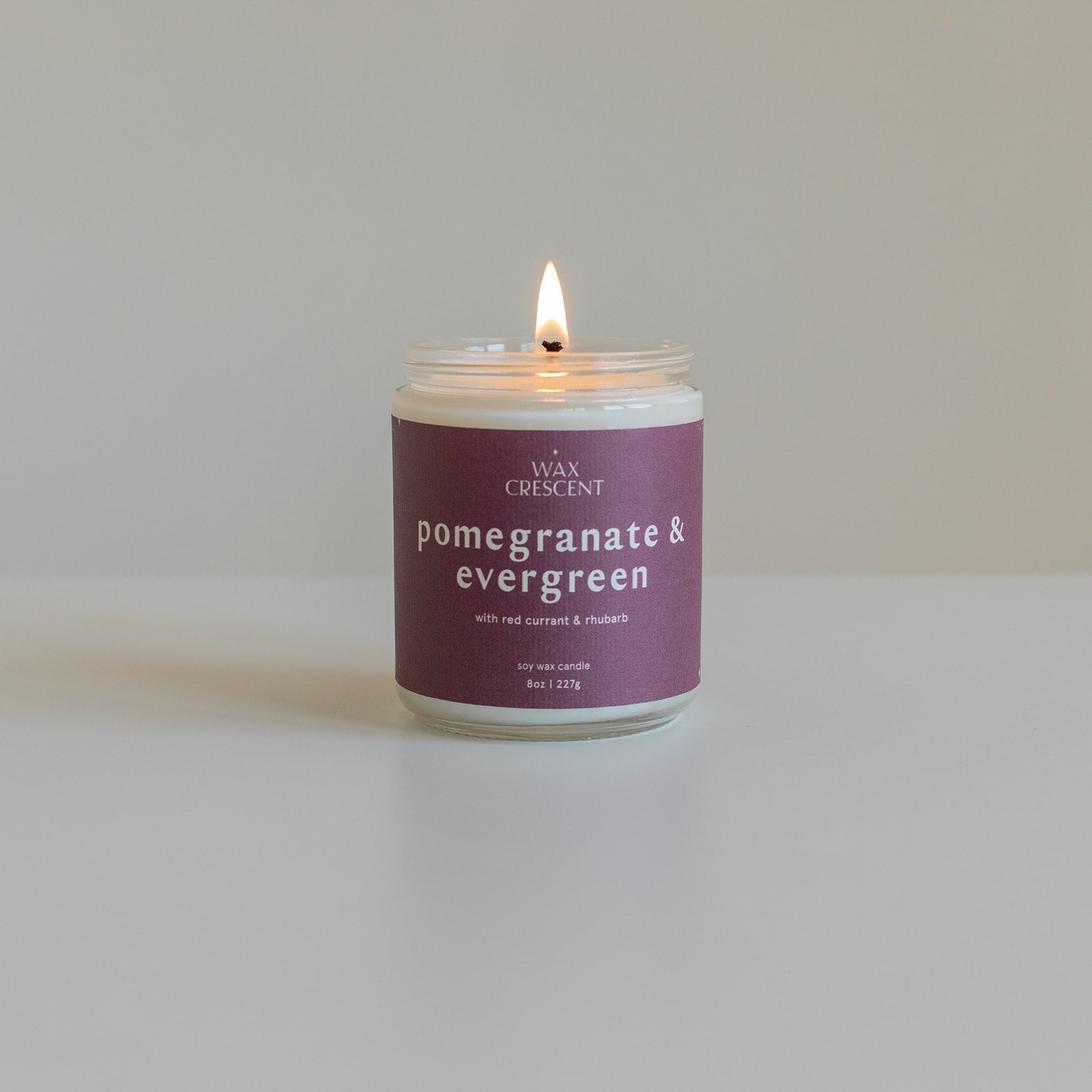 A lit pomegranate & evergreen soy wax candle from Wax Crescent, featuring a deep purple label, placed on a clean white surface. The candle's label highlights its natural ingredients and rich fruity and evergreen scent