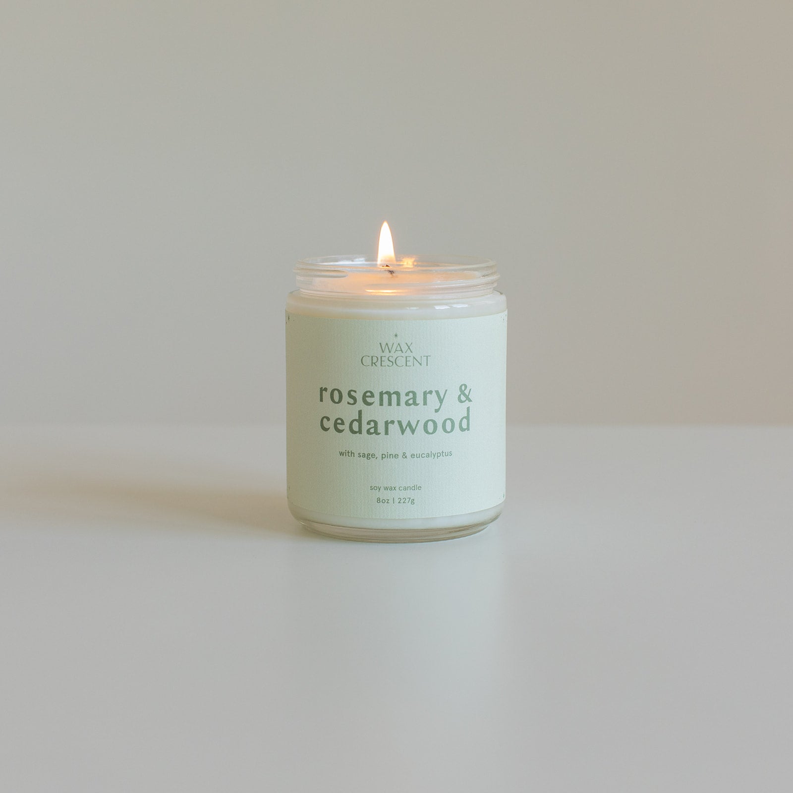 A lit rosemary & cedarwood soy wax candle from Wax Crescent, with a minimalist label, placed on a clean white surface. The candle's label highlights its natural ingredients, offering a calming and earthy scent