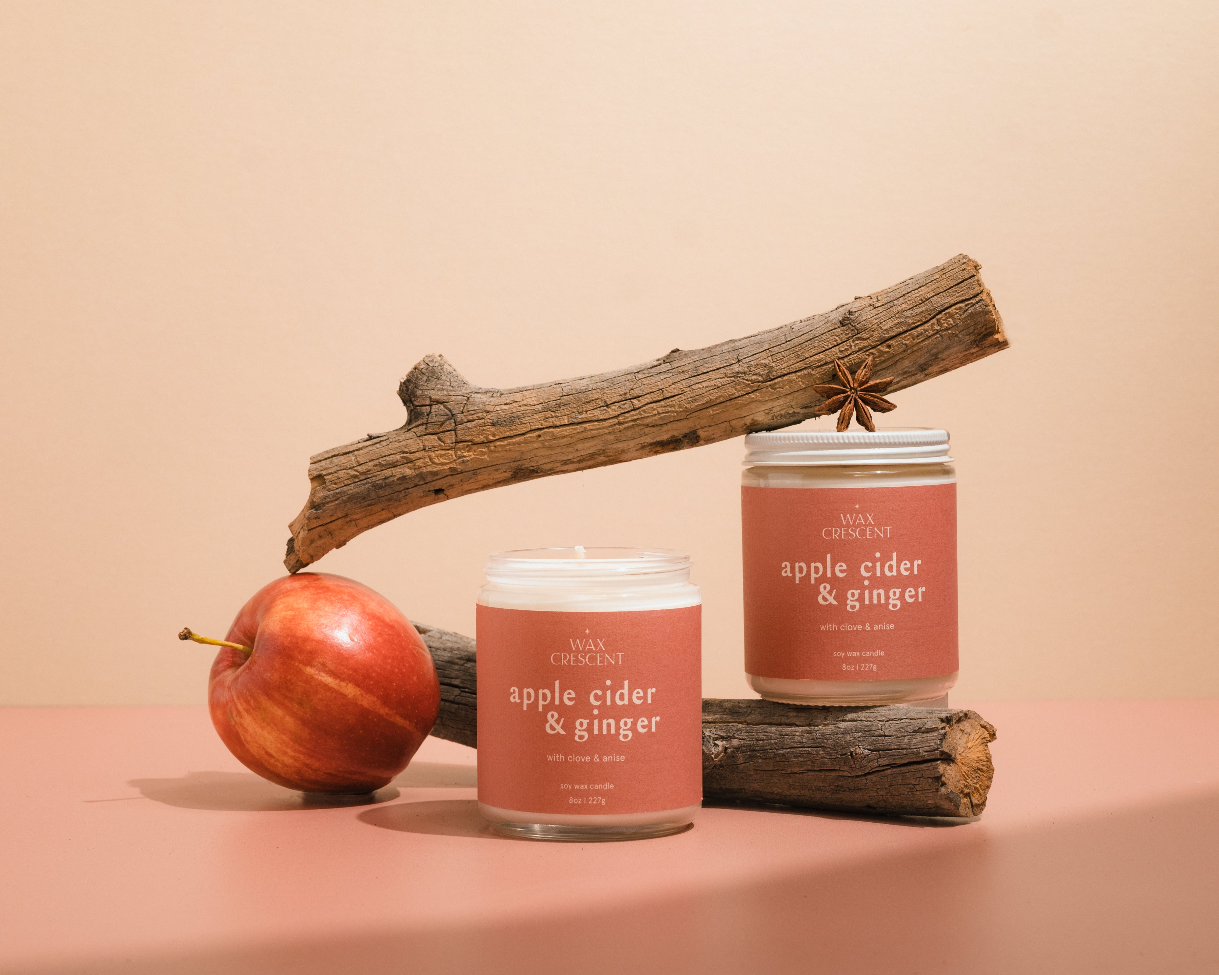 Two Apple Cider & Ginger luxury soy wax candles on driftwood, featuring anise seeds, highlighting their elegant design and warm colors, perfect for creating a cozy autumn atmosphere.