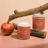 Two Apple Cider & Ginger luxury soy wax candles alongside a fresh apple, highlighting their warm colors and inviting design, perfect for creating a cozy autumn ambiance.