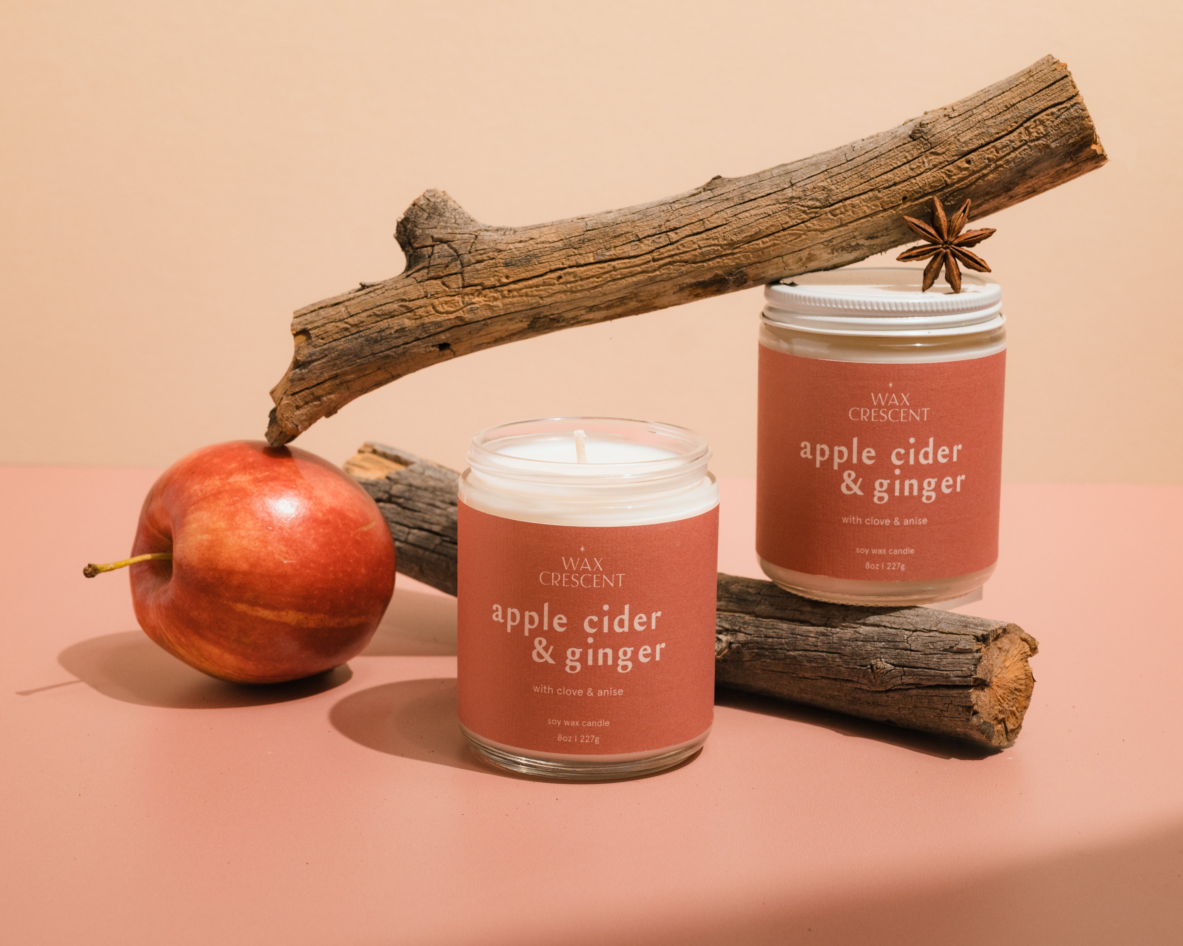 Two Apple Cider & Ginger luxury soy wax candles alongside a fresh apple, highlighting their warm colors and inviting design, perfect for creating a cozy autumn ambiance.