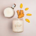 A flat lay of a Cardamom & Cream candle next to a small metal cup filled with milk and a piece of fresh ginger, with yellow ginger slices scattered around, arranged in a creative pattern on a light background.