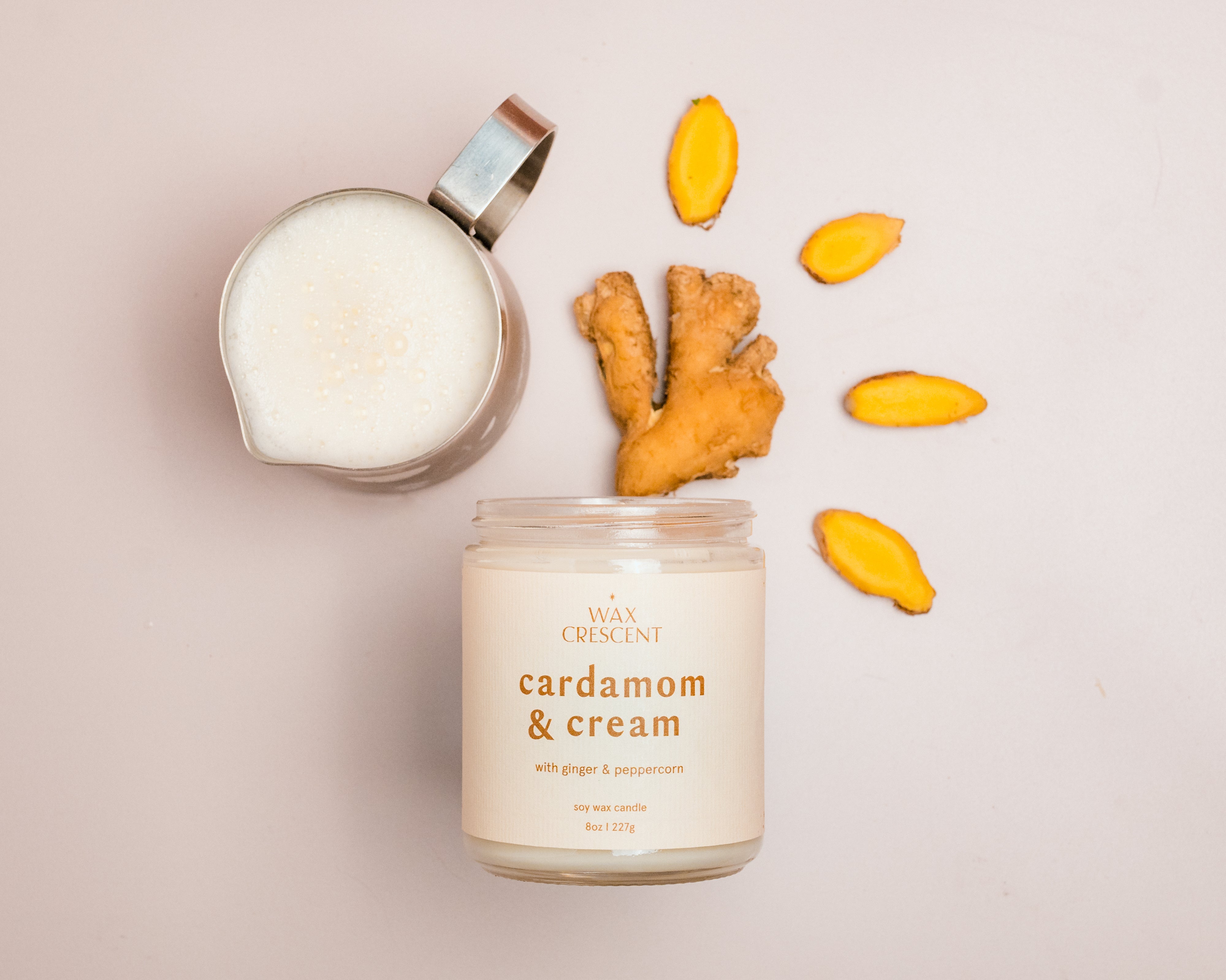 A flat lay of a Cardamom & Cream candle next to a small metal cup filled with milk and a piece of fresh ginger, with yellow ginger slices scattered around, arranged in a creative pattern on a light background.