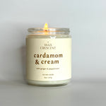 A lit Cardamom & Cream candle on a clean, minimalist surface, giving off a warm, cozy glow.
