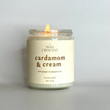 A lit Cardamom & Cream candle on a clean, minimalist surface, giving off a warm, cozy glow.