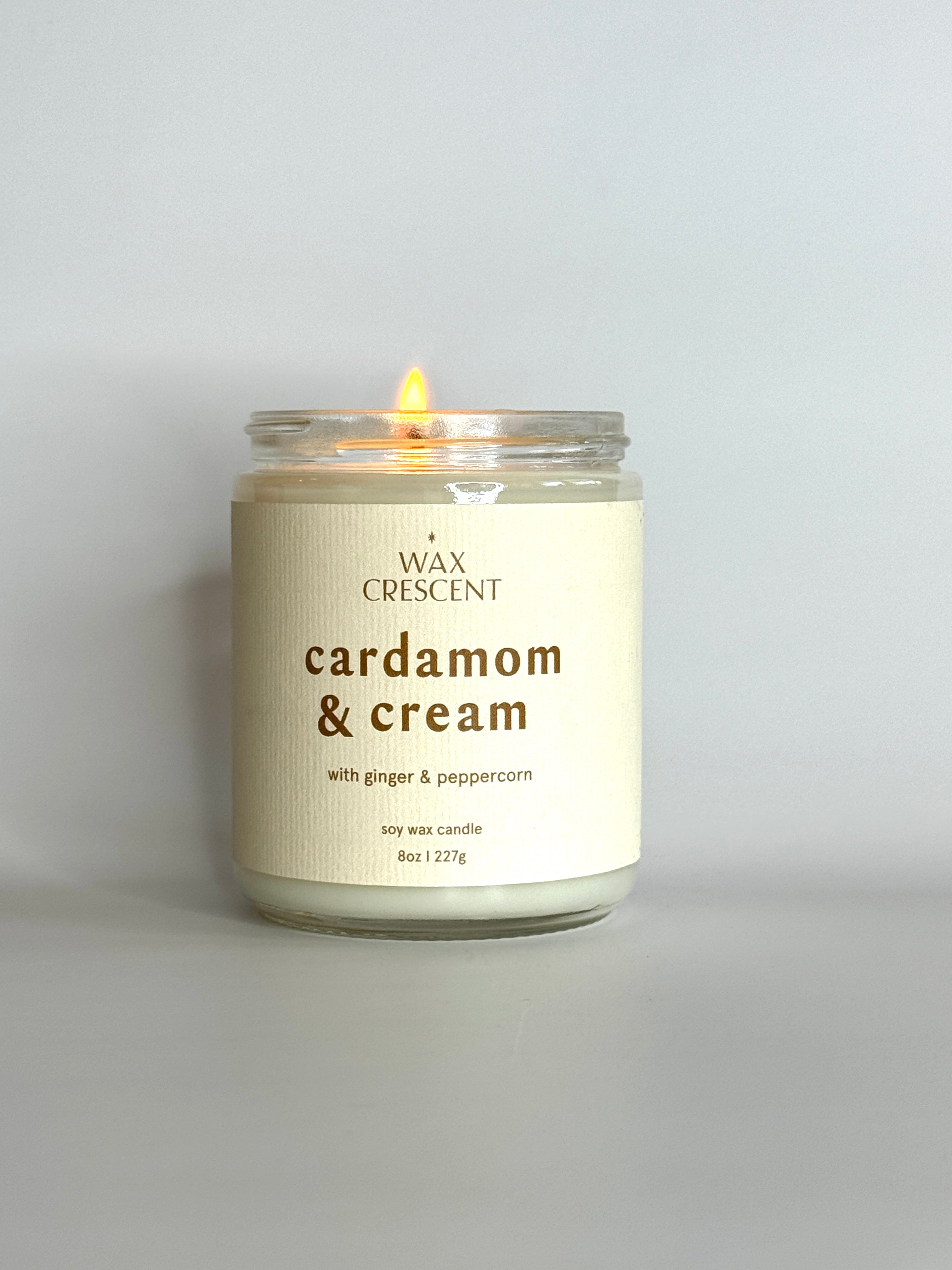A lit Cardamom & Cream candle on a clean, minimalist surface, giving off a warm, cozy glow.