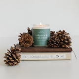 Tobacco & Teakwood candle displayed on a rustic wood slab, surrounded by pine cones, creating a warm and inviting autumn ambiance.
