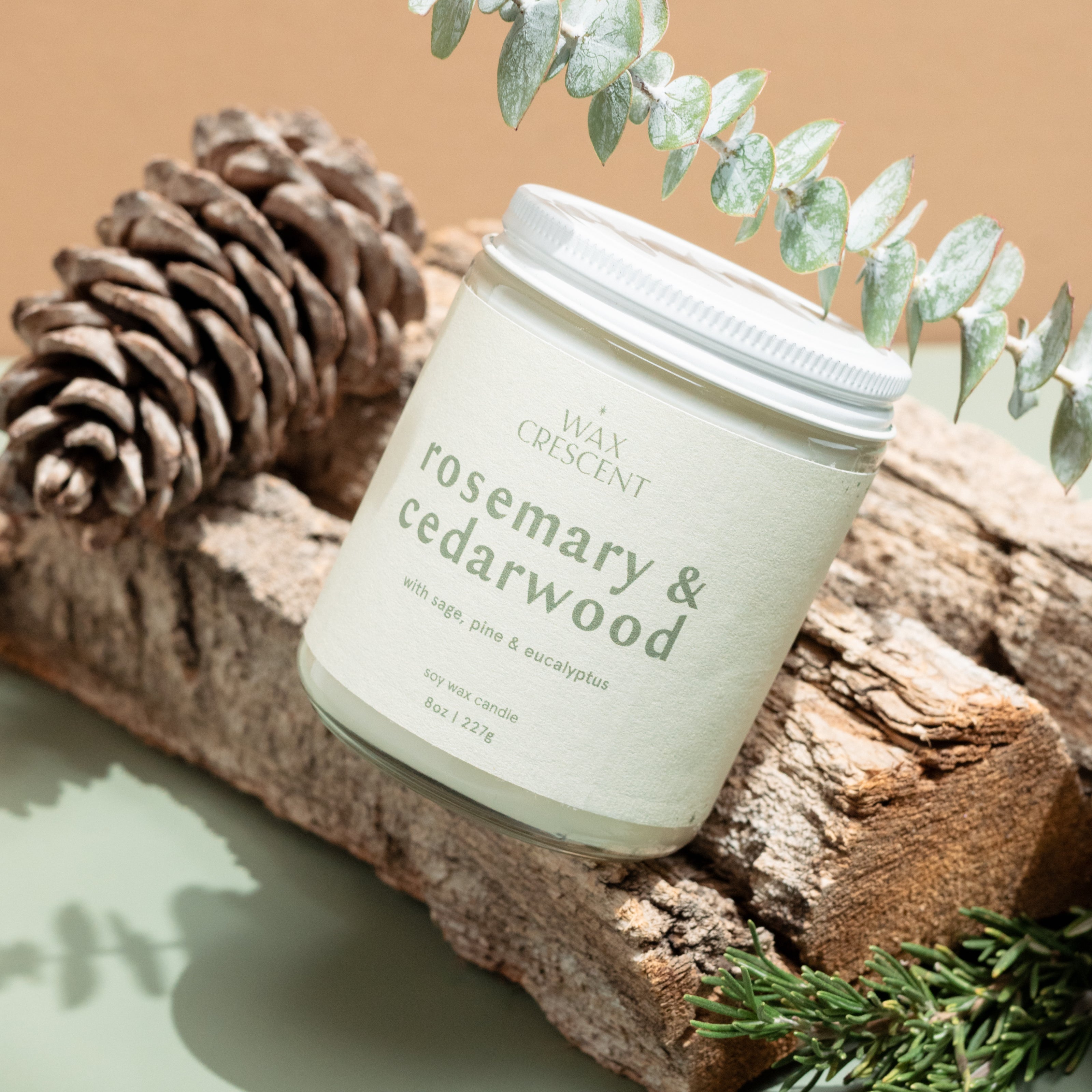 Wax Crescent Rosemary & Cedarwood soy candle placed on a rustic wood log, surrounded by pinecones, fresh rosemary sprigs, and eucalyptus. A nature-inspired, earthy aesthetic.