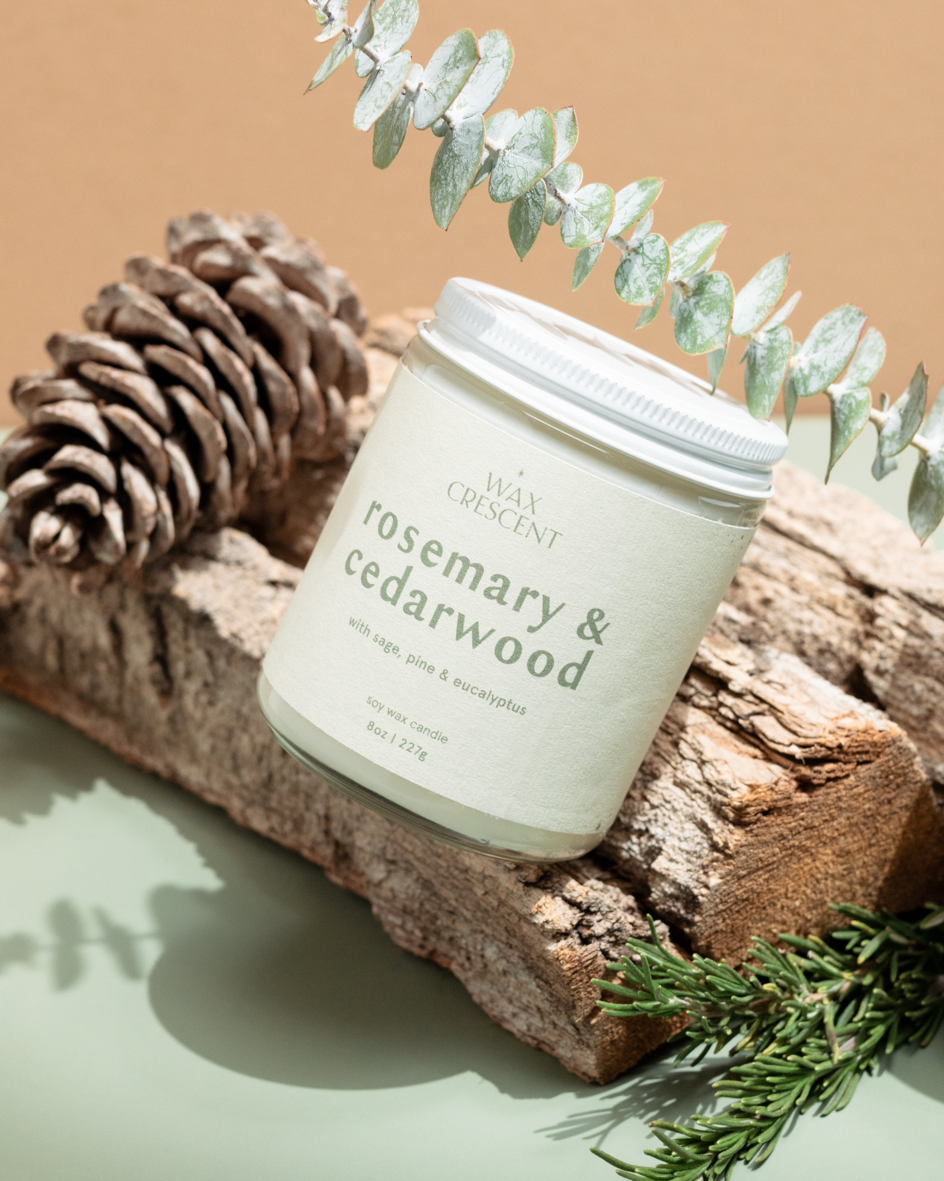 Wax Crescent Rosemary & Cedarwood soy candle placed on a rustic wood log, surrounded by pinecones, fresh rosemary sprigs, and eucalyptus. A nature-inspired, earthy aesthetic.