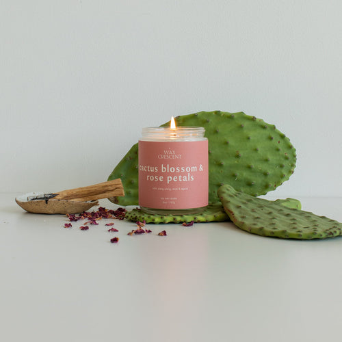cactus blossom & rose petals candle with pink label lit on a table surrounded by dried rose petals and cactus leaves. 