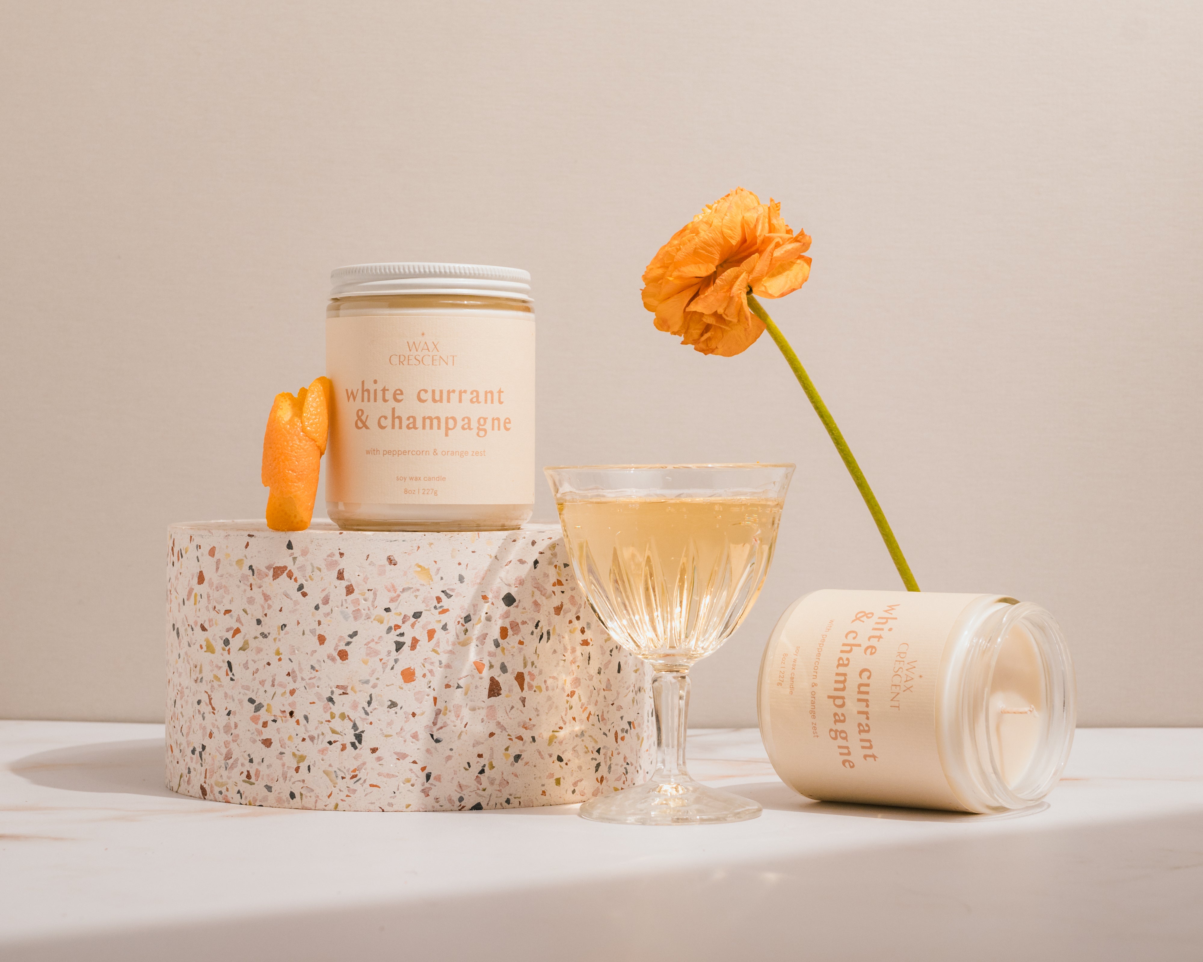 Non-toxic soy candles for creating a cozy home next to orange flowers a glass of sparkling wine and stacked on terrazo blocks