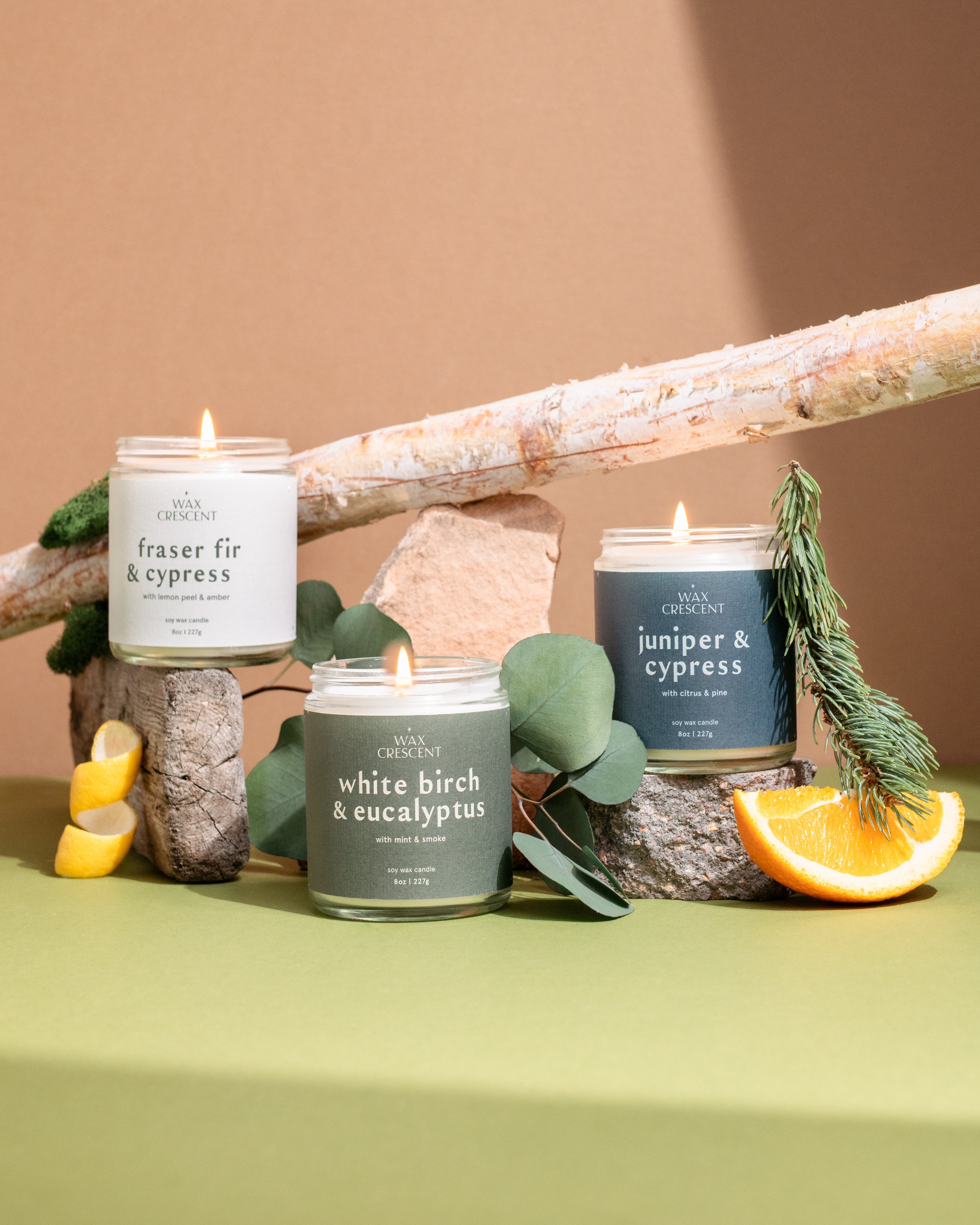 Wax Crescent winter candles including Cranberry & Balsam, Oakmoss & Evergreen, and Fraser Fir & Cypress, styled with pinecones and evergreen branches to evoke a cozy, seasonal winter atmosphere
