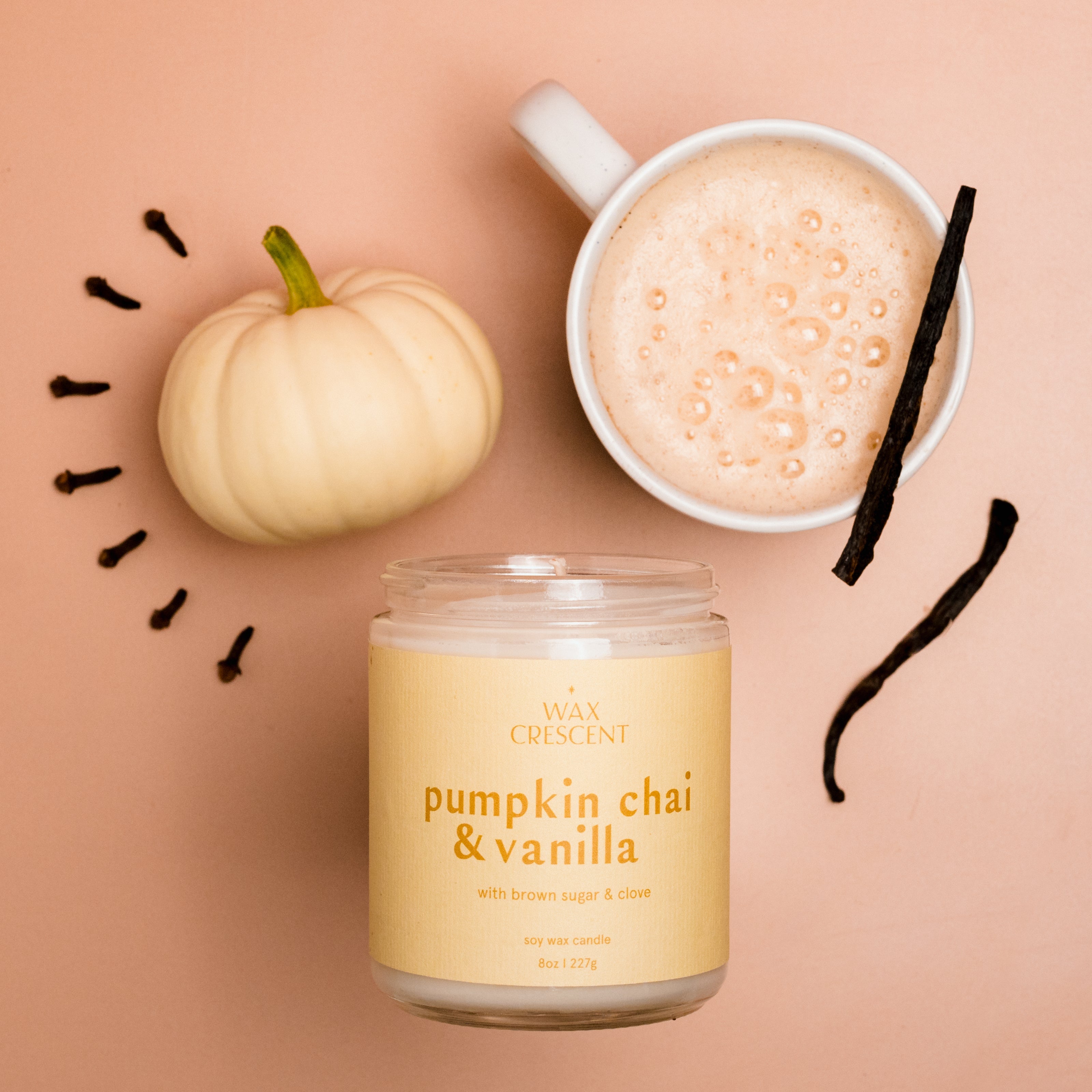 Pumpkin Chai & Vanilla soy candle styled with a creamy chai latte, vanilla bean, and a white pumpkin, evoking cozy fall vibes and warm autumn scents.