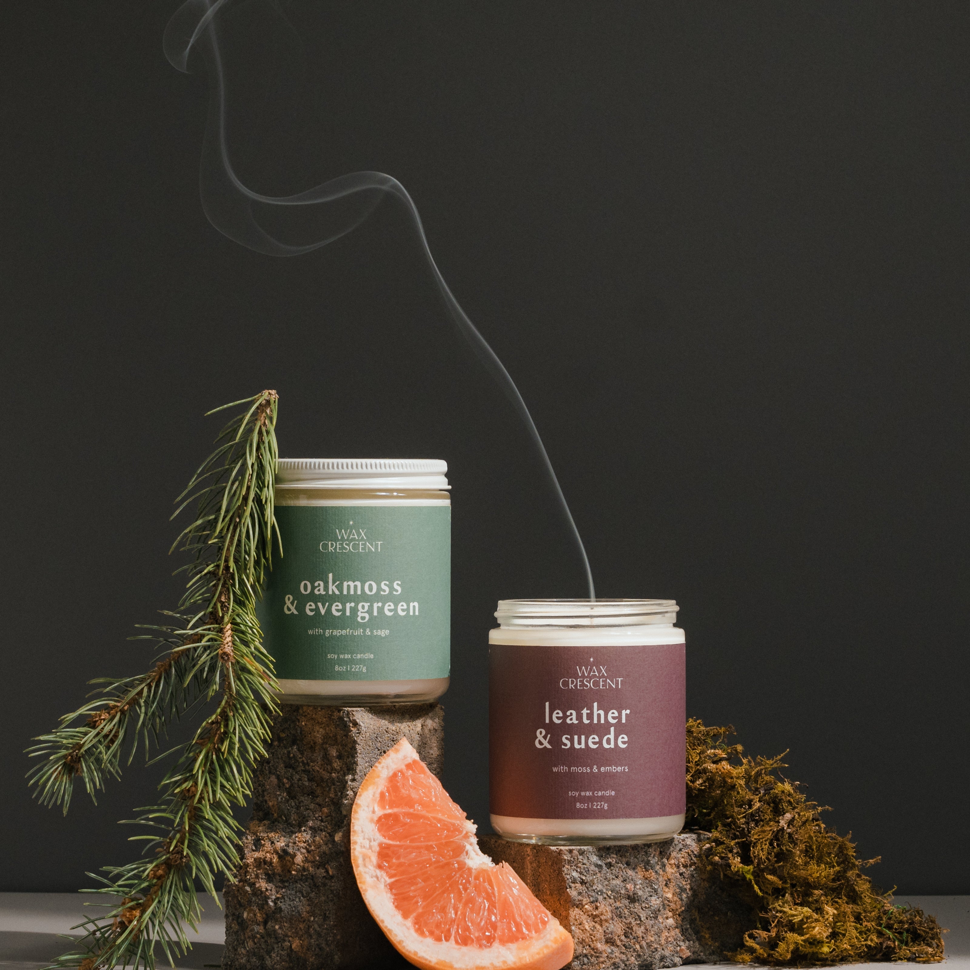 Two Wax Crescent soy wax candles, Oakmoss & Evergreen and Leather & Suede, displayed with fresh greenery and a grapefruit slice. The candles are part of a monthly subscription box offering curated seasonal scents