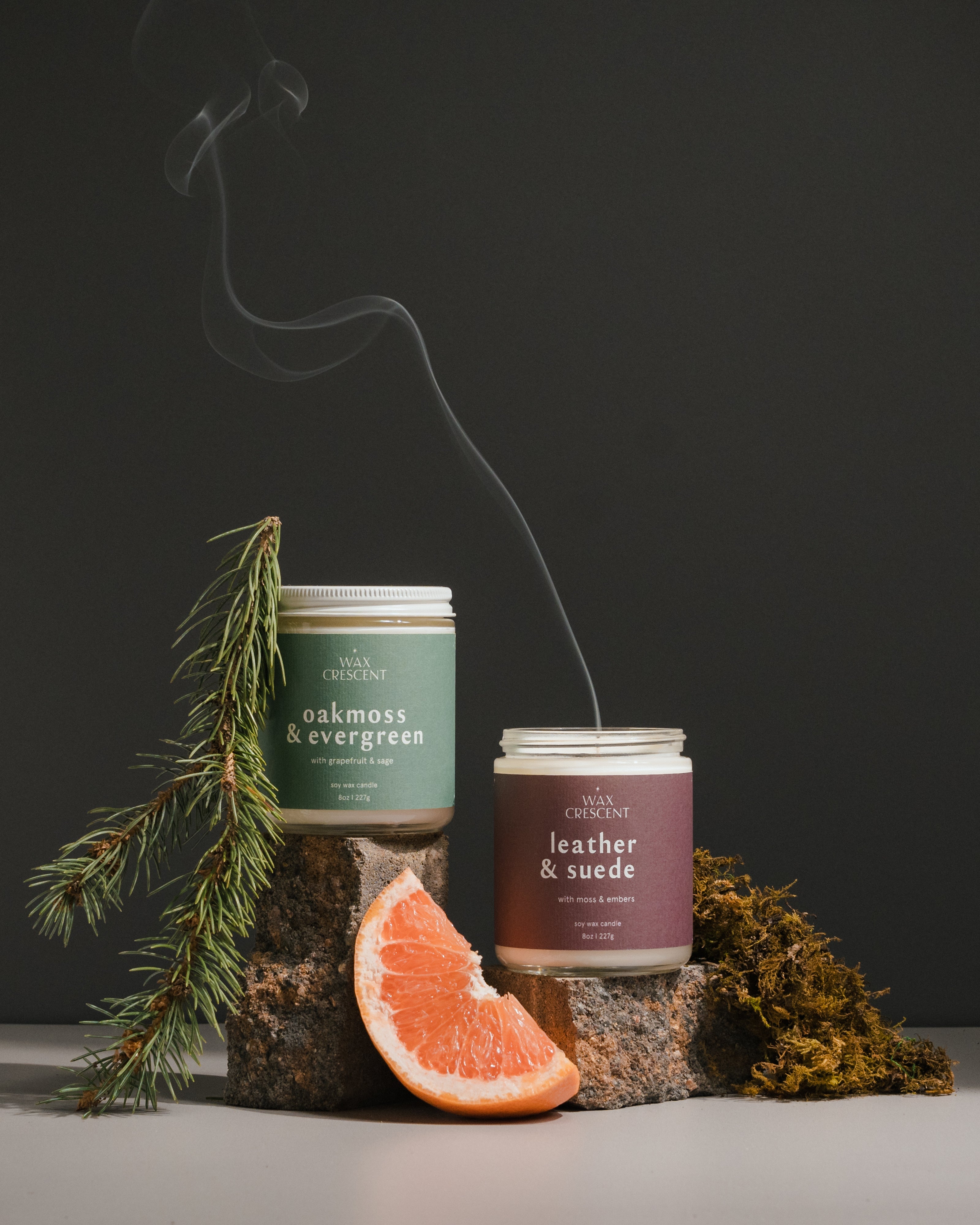 Two Wax Crescent soy wax candles, Oakmoss & Evergreen and Leather & Suede, displayed with fresh greenery and a grapefruit slice. The candles are part of a monthly subscription box offering curated seasonal scents