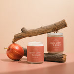 Two Apple Cider & Ginger luxury soy wax candles on driftwood, featuring anise seeds, highlighting their elegant design and warm colors, perfect for creating a cozy autumn atmosphere.