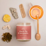 Flat lay of Black Fig & Bergamot candle with mauve label, surrounded by honey, lemon, ginger, and tea leaves