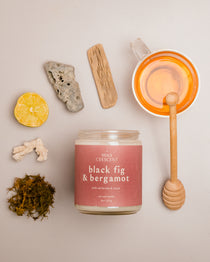 Flat lay of Black Fig & Bergamot candle with mauve label, surrounded by honey, lemon, ginger, and tea leaves