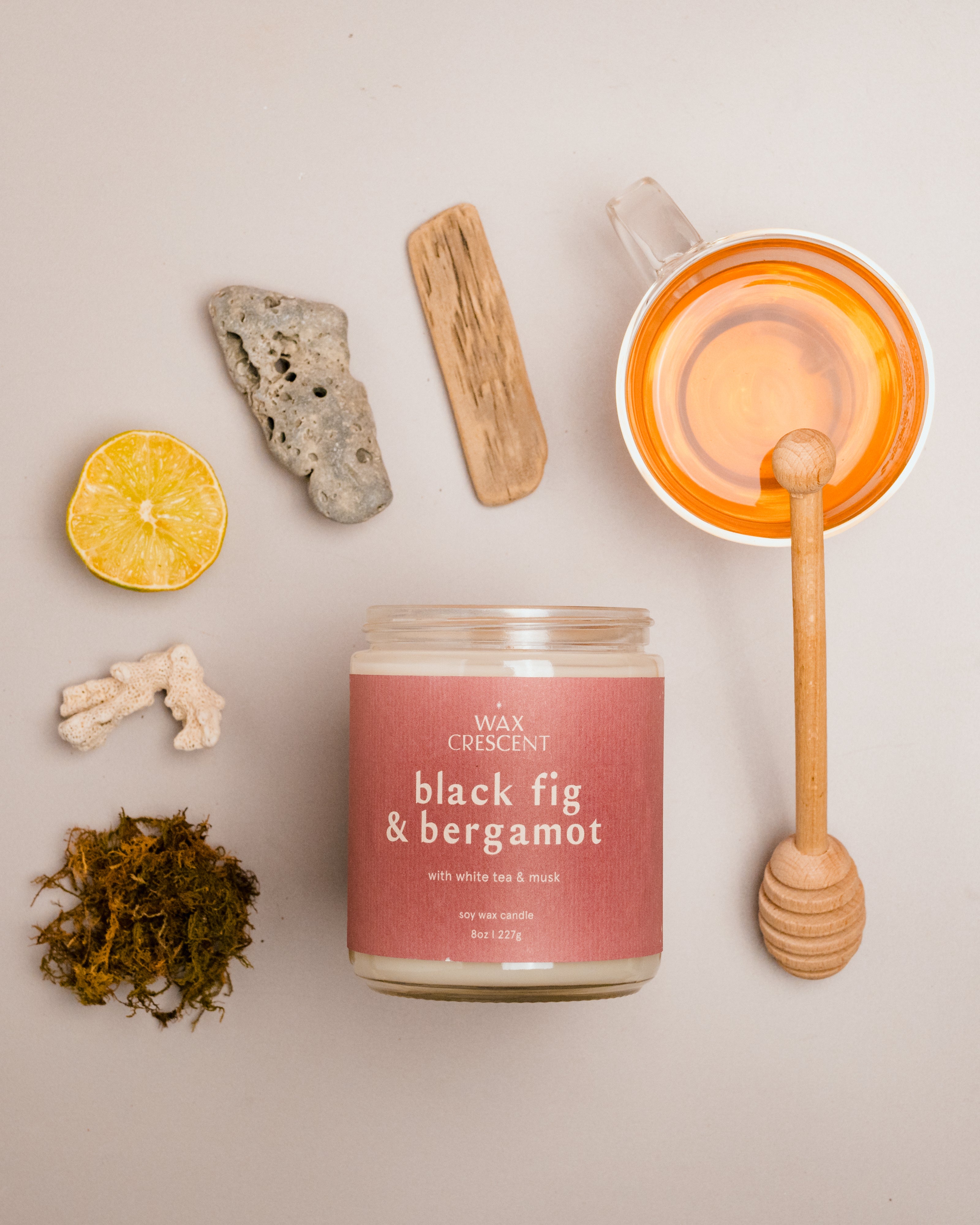 Flat lay of Black Fig & Bergamot candle with mauve label, surrounded by honey, lemon, ginger, and tea leaves