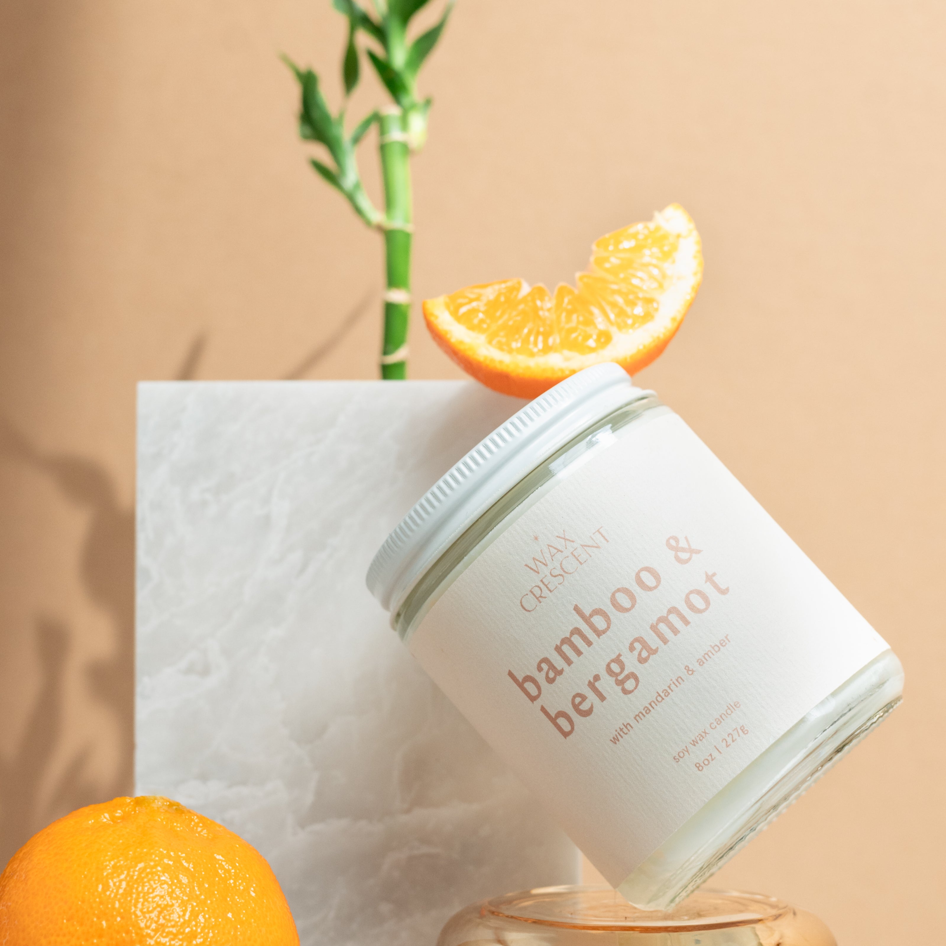 close up of Wax Crescent Bamboo & Bergamot candle styled with fresh oranges, bamboo stems, and natural greenery against a minimalist beige backdrop. A refreshing, spa-like ambiance