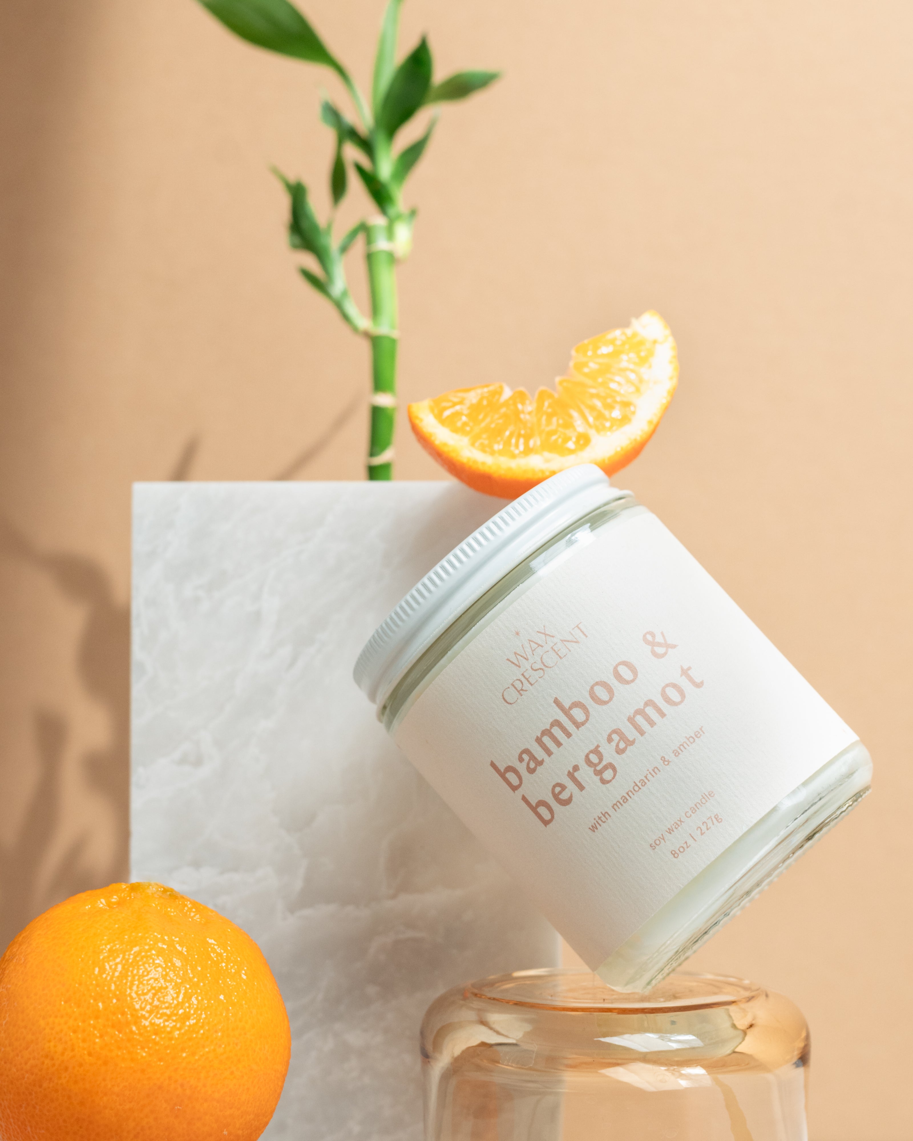 close up of Wax Crescent Bamboo & Bergamot candle styled with fresh oranges, bamboo stems, and natural greenery against a minimalist beige backdrop. A refreshing, spa-like ambiance