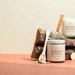 Two Cardamom & Cream luxury soy candles resting on a wooden log with cinnamon sticks and cardamom pods — a warm, creamy blend of spice and vanilla hand-poured by Wax Crescent, the perfect non-toxic holiday candle for cozy winter gifting