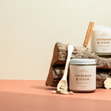 Two Cardamom & Cream luxury soy candles resting on a wooden log with cinnamon sticks and cardamom pods — a warm, creamy blend of spice and vanilla hand-poured by Wax Crescent, the perfect non-toxic holiday candle for cozy winter gifting
