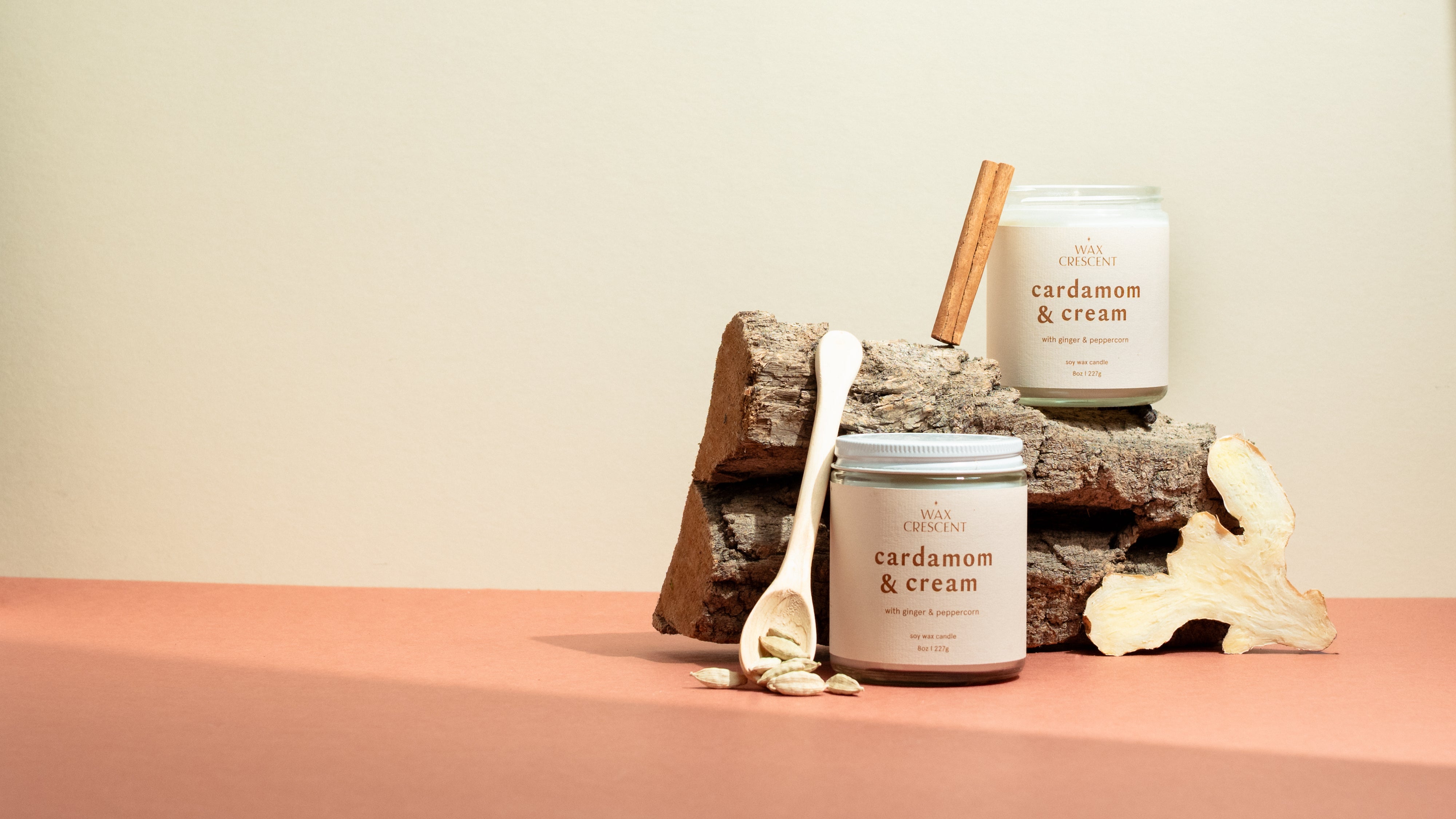 Two Cardamom & Cream luxury soy candles resting on a wooden log with cinnamon sticks and cardamom pods — a warm, creamy blend of spice and vanilla hand-poured by Wax Crescent, the perfect non-toxic holiday candle for cozy winter gifting