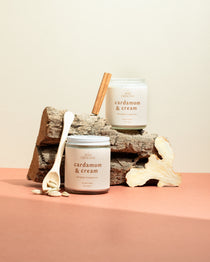 Two Cardamom & Cream soy candles on a wooden log surrounded by cinnamon sticks and cardamom pods — a warm, spiced fragrance hand-poured by Wax Crescent for cozy winter nights.