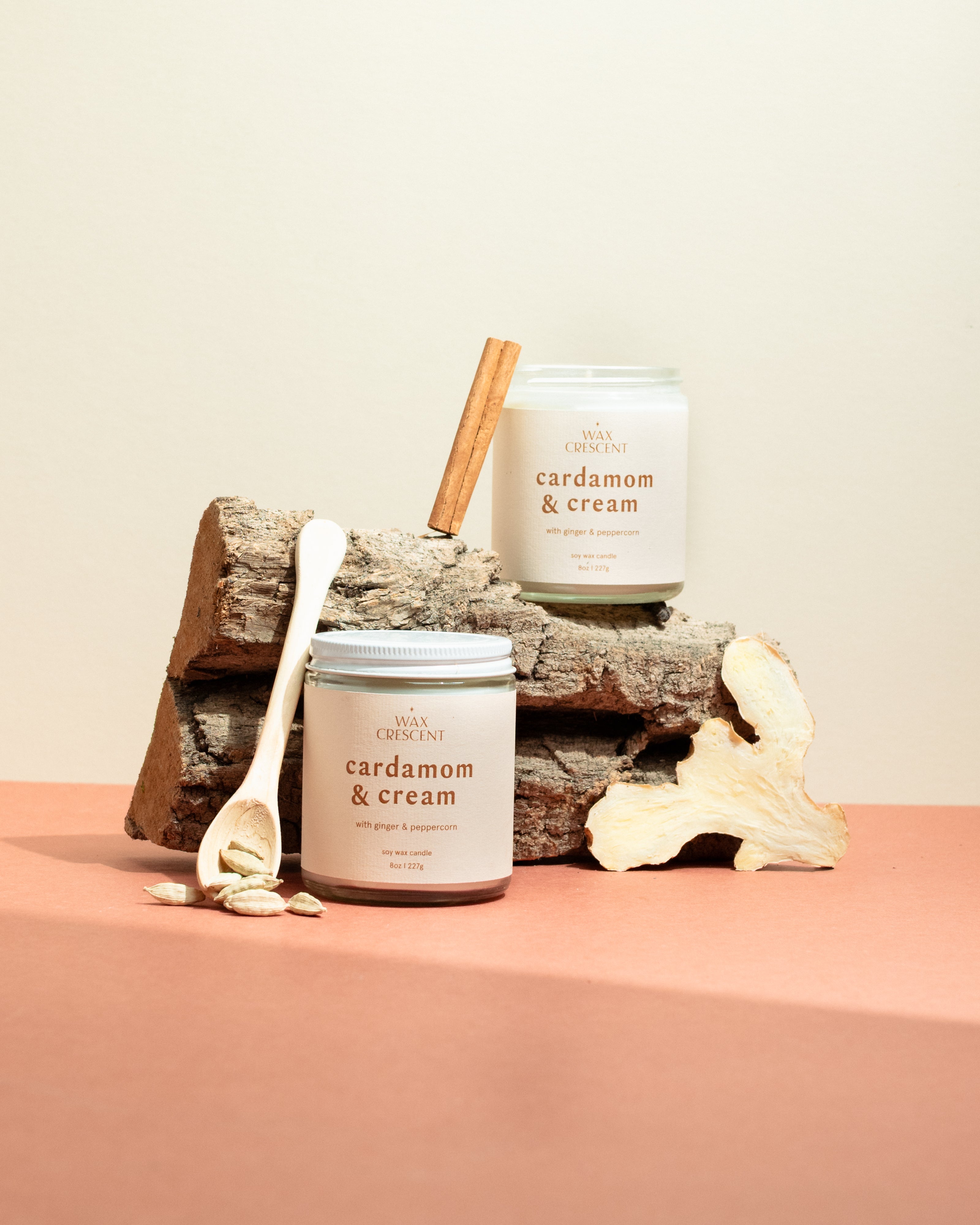 Two Cardamom & Cream soy candles on a wooden log surrounded by cinnamon sticks and cardamom pods — a warm, spiced fragrance hand-poured by Wax Crescent for cozy winter nights.
