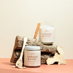 Pair of Cardamom & Cream candles on a wooden log with cinnamon sticks and cardamom pods — luxury soy candles from Wax Crescent blending creamy vanilla warmth with aromatic spice