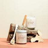 Pair of Cardamom & Cream candles on a wooden log with cinnamon sticks and cardamom pods — luxury soy candles from Wax Crescent blending creamy vanilla warmth with aromatic spice