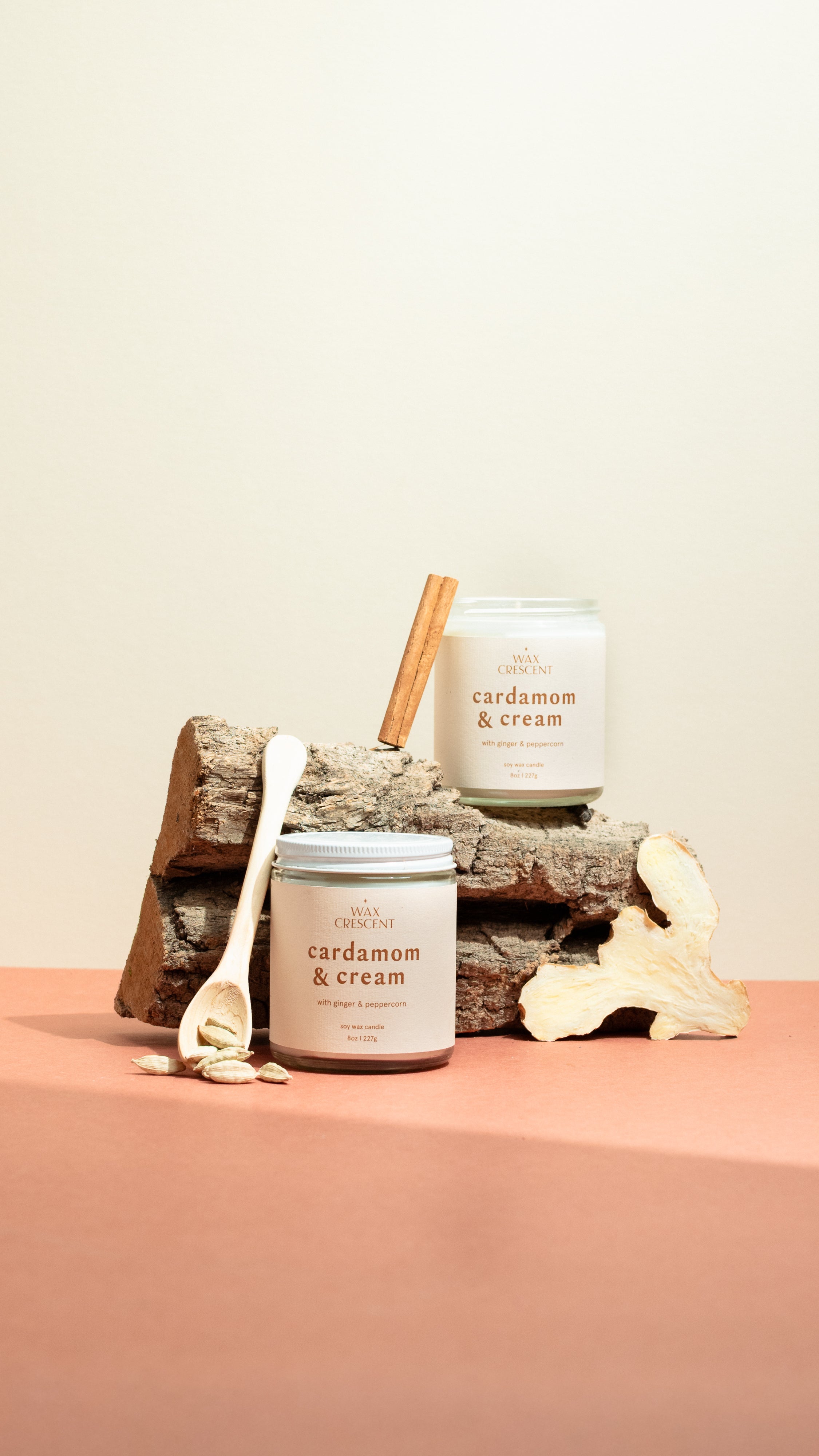Pair of Cardamom & Cream candles on a wooden log with cinnamon sticks and cardamom pods — luxury soy candles from Wax Crescent blending creamy vanilla warmth with aromatic spice