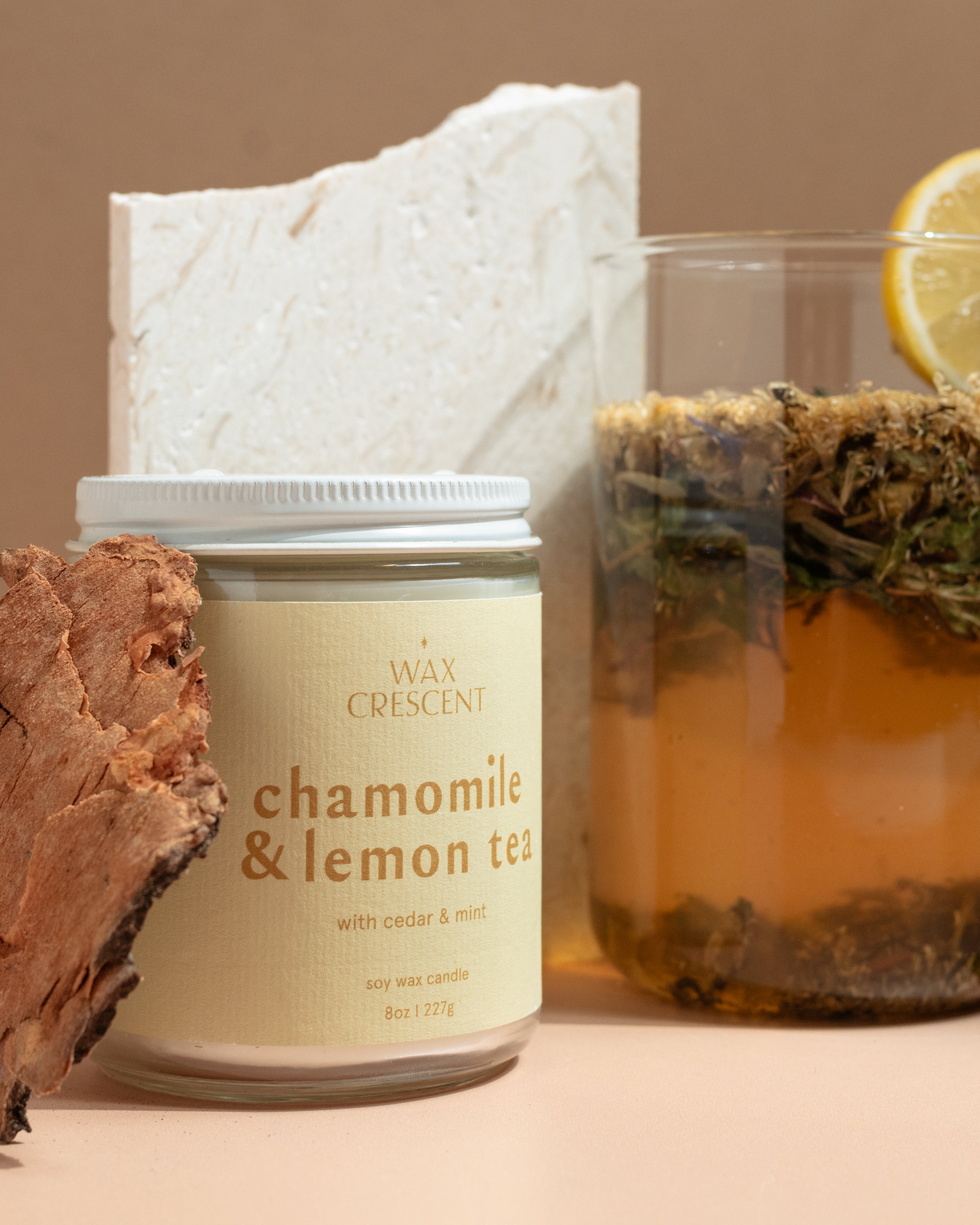 clos e up side angle of Wax Crescent Chamomile & Lemon Tea candle styled with a glass of herbal tea, lemon slice, mint leaves, and natural bark against a soft beige background 