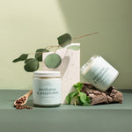Eucalyptus & Peppermint holiday candle duo on a green surface with eucalyptus leaves and a pine cone — fresh, festive aroma hand-poured by Wax Crescent for the winter season.