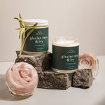 A close-up of two Glacier Rose & Ivy candles by Wax Crescent, styled with natural elements like a pink rose and sage on textured blocks, showcasing a minimalist aesthetic