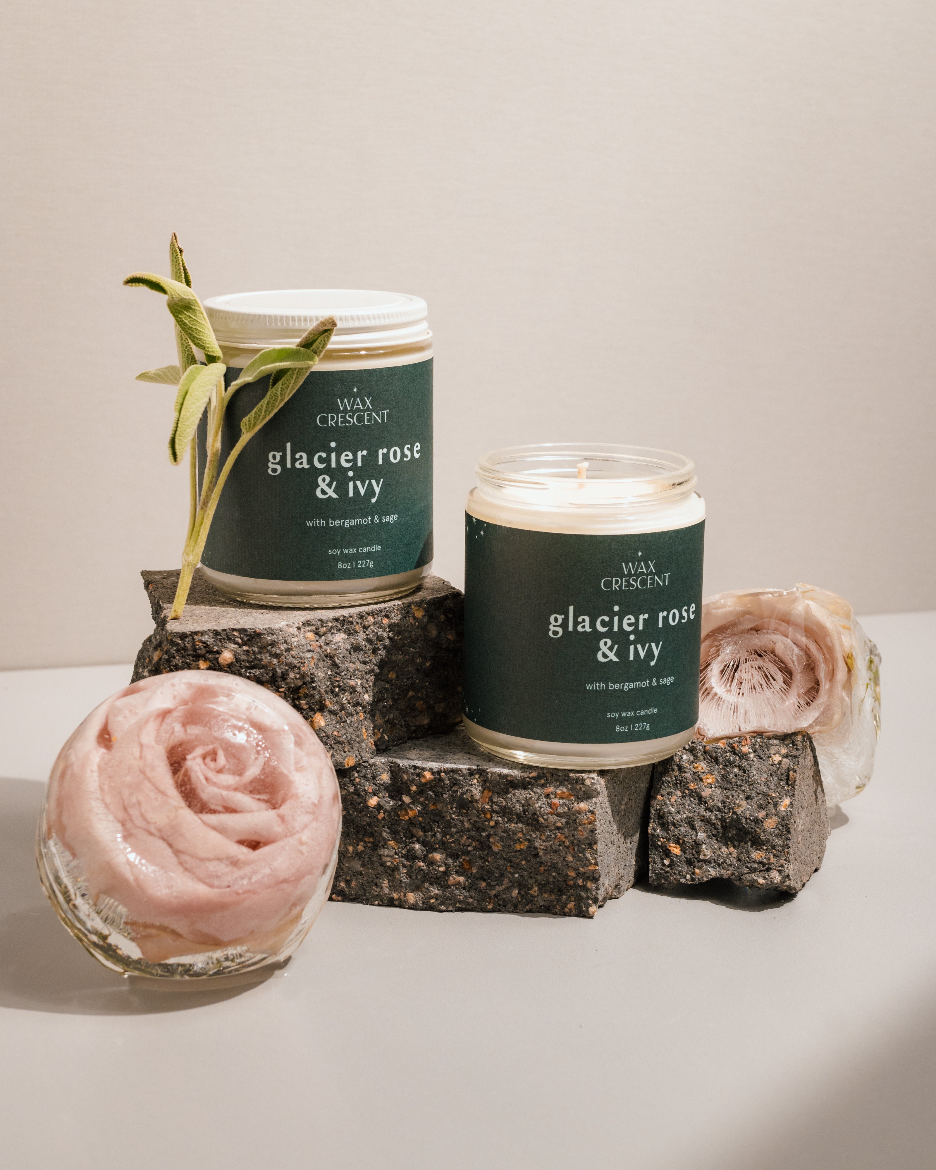 A close-up of two Glacier Rose & Ivy candles by Wax Crescent, styled with natural elements like a pink rose and sage on textured blocks, showcasing a minimalist aesthetic