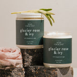 A close-up of two Glacier Rose & Ivy candles by Wax Crescent, styled with natural elements like a pink rose and sage on textured blocks, showcasing a minimalist aesthetic