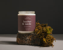 Leather & Suede luxury soy candle styled on rustic cement block with moss, perfect for creating a masculine and earthy ambiance