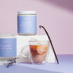 Two lavender-scented candles and a glass of iced coffee on a purple surface with a vanilla bean. Hand poured 