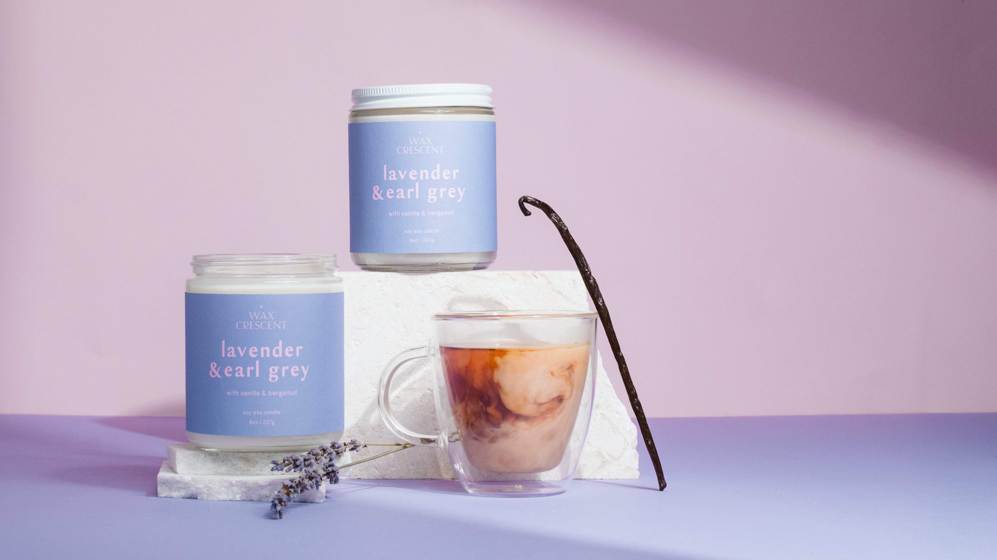 Two lavender-scented candles and a glass of iced coffee on a purple surface with a vanilla bean. Hand poured 