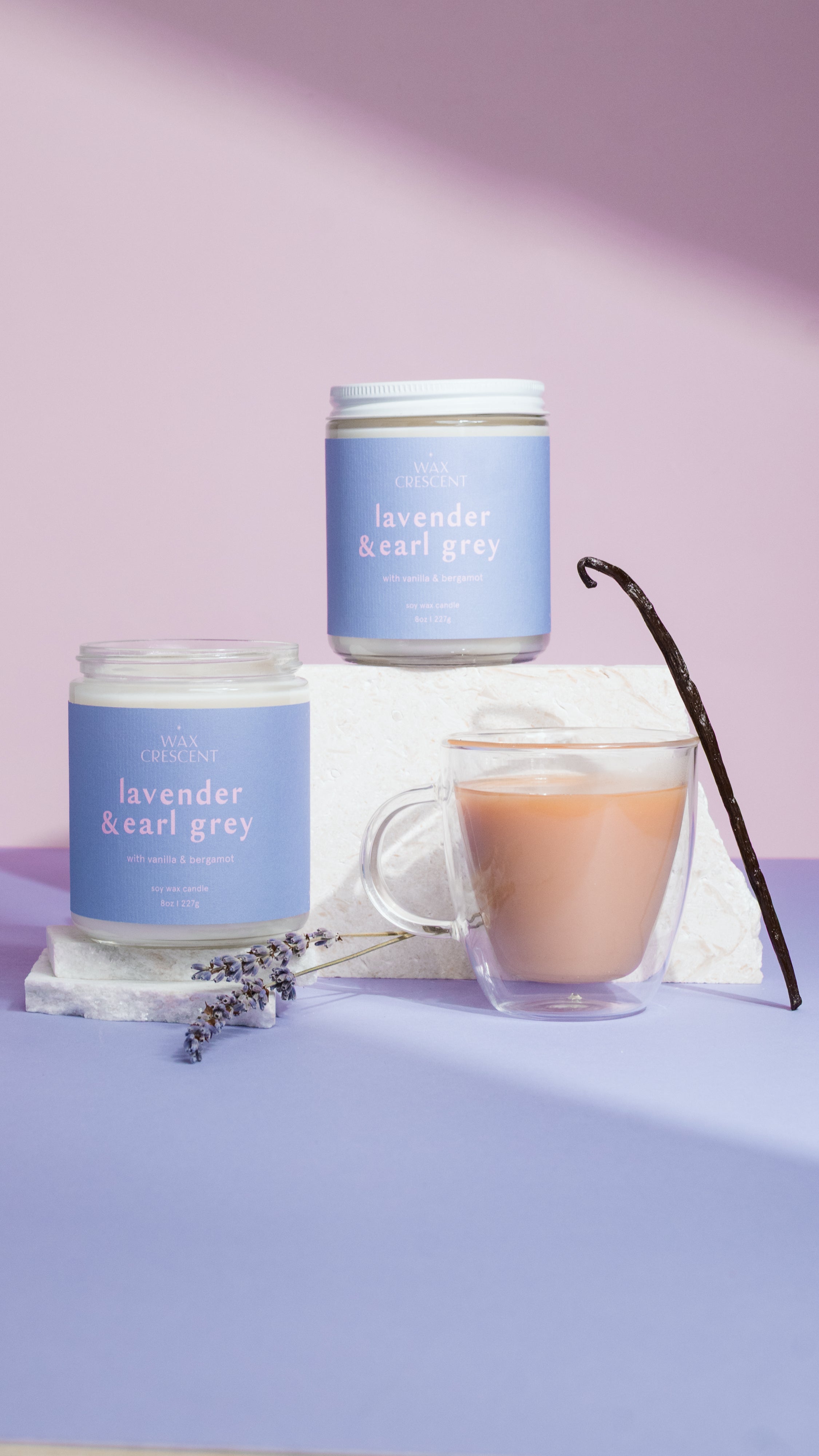 Two lavender-scented candles and a cup of tea on a purple surface with a light purple background.
