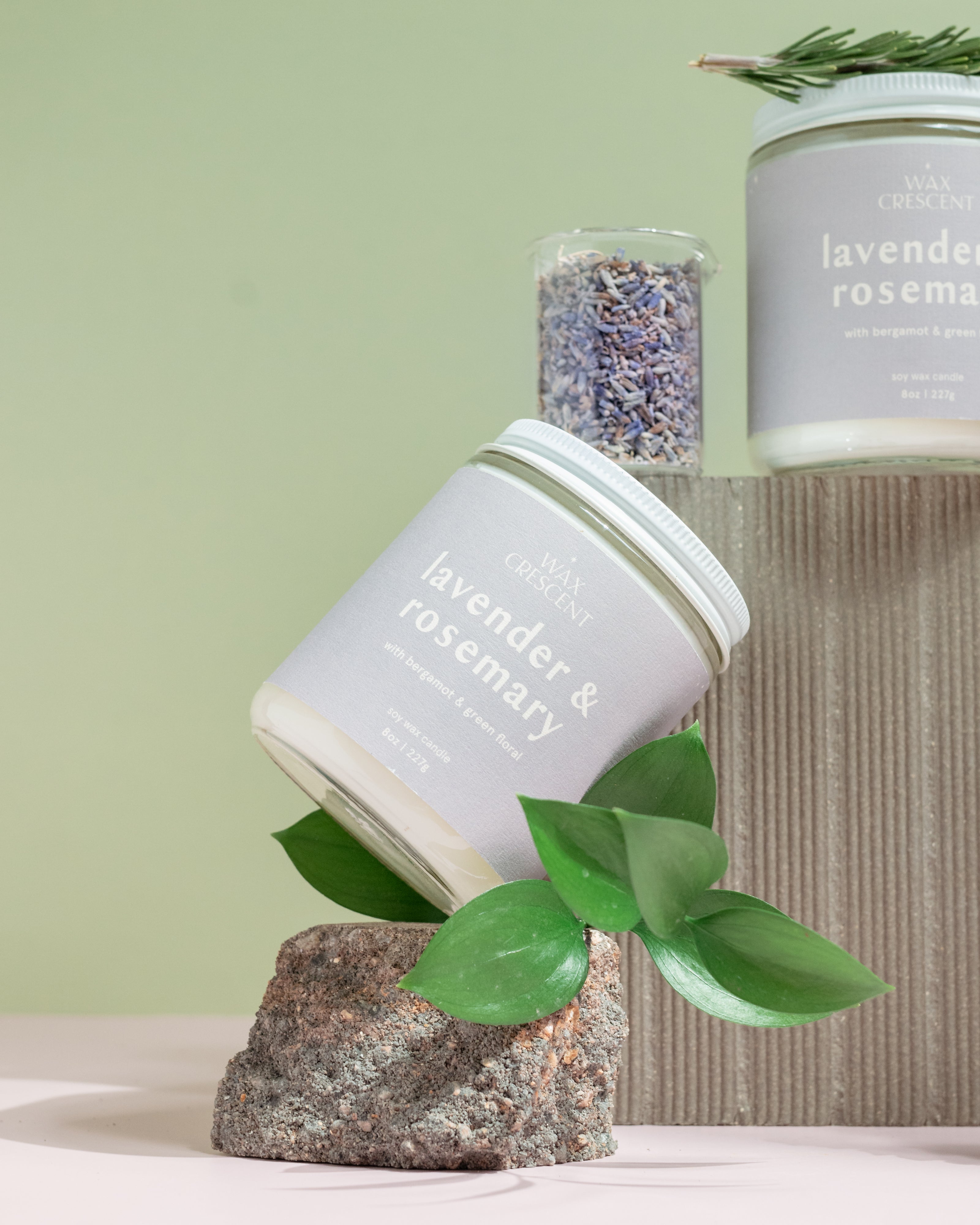 close up of Wax Crescent Lavender & Rosemary candle styled with natural elements like lavender buds, rosemary sprigs, and amethyst crystal, set against a calming pastel backdrop