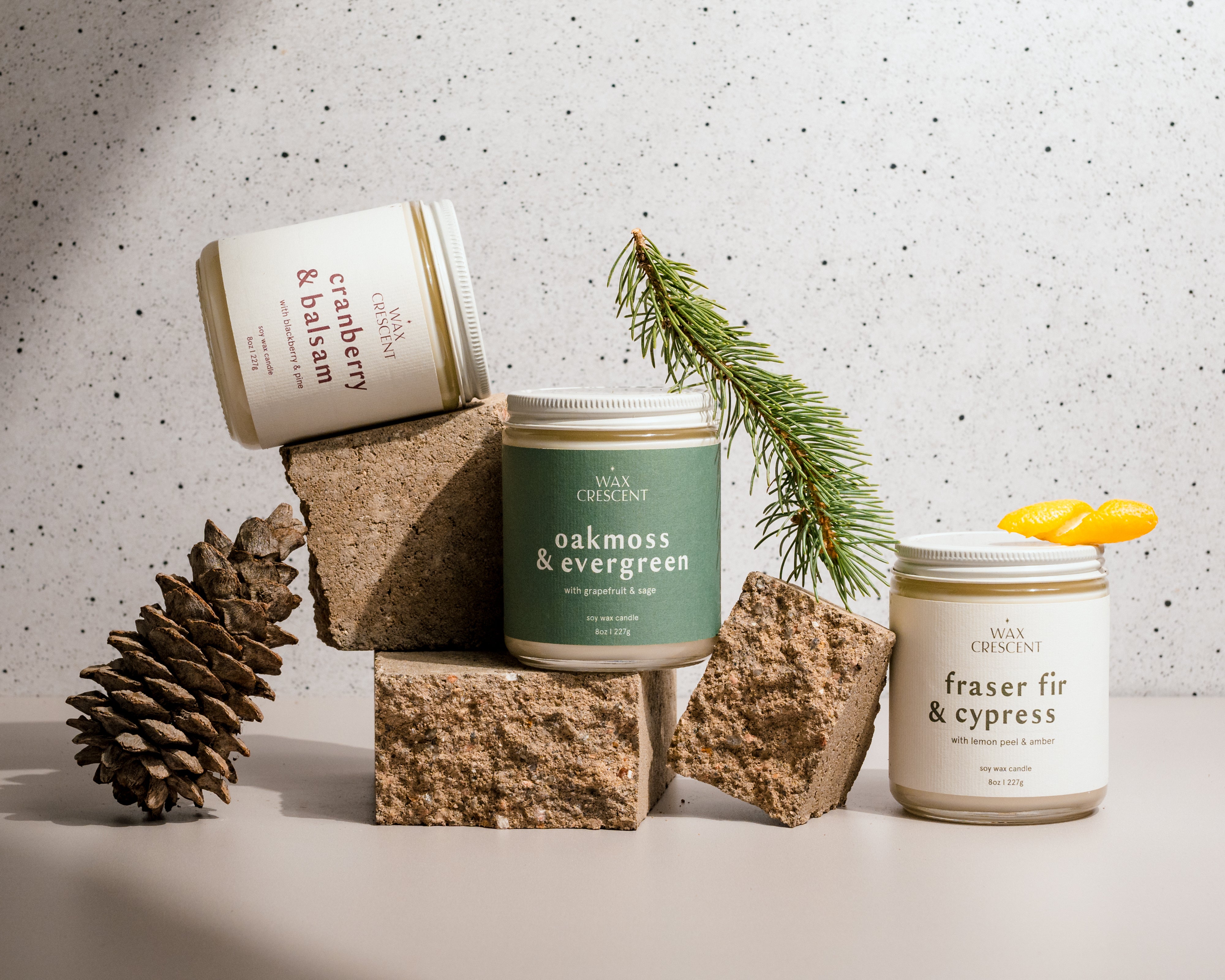 Three winter candles—Cranberry & Balsam, Oakmoss & Evergreen, and Fraser Fir & Cypress—playfully displayed among pine cones, lemon slices, and evergreen sprigs on natural rocks, capturing a festive and nature-inspired setting