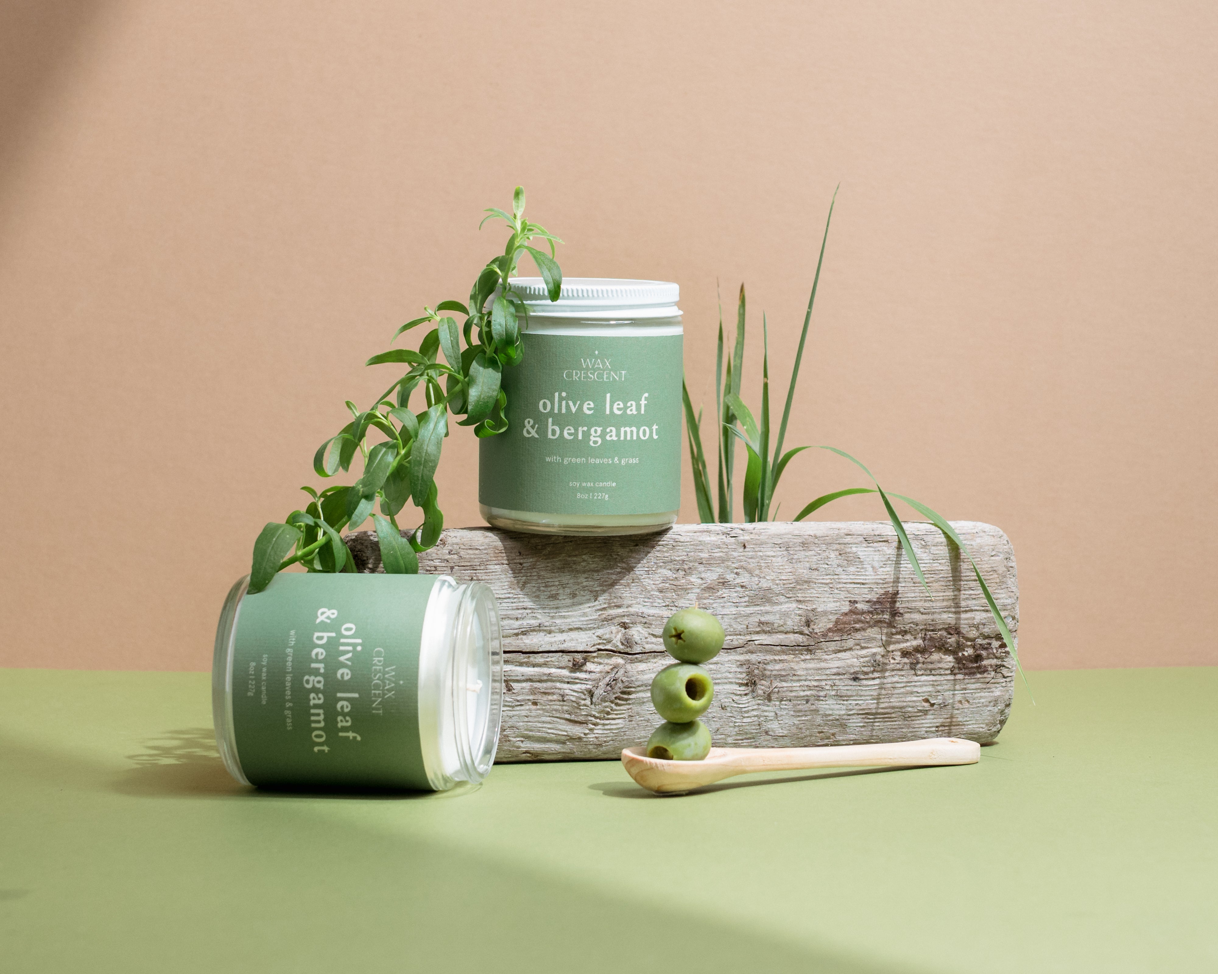 Pair of Olive Leaf & Bergamot candles on a neutral backdrop surrounded by olives and leaves — small-batch soy candles from Wax Crescent that bring fresh, botanical warmth to any space.