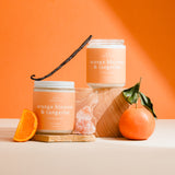 Two candles labeled 'Orange Blossom & Tangerine' with an orange, vanilla bean, and leaf on a peach background.