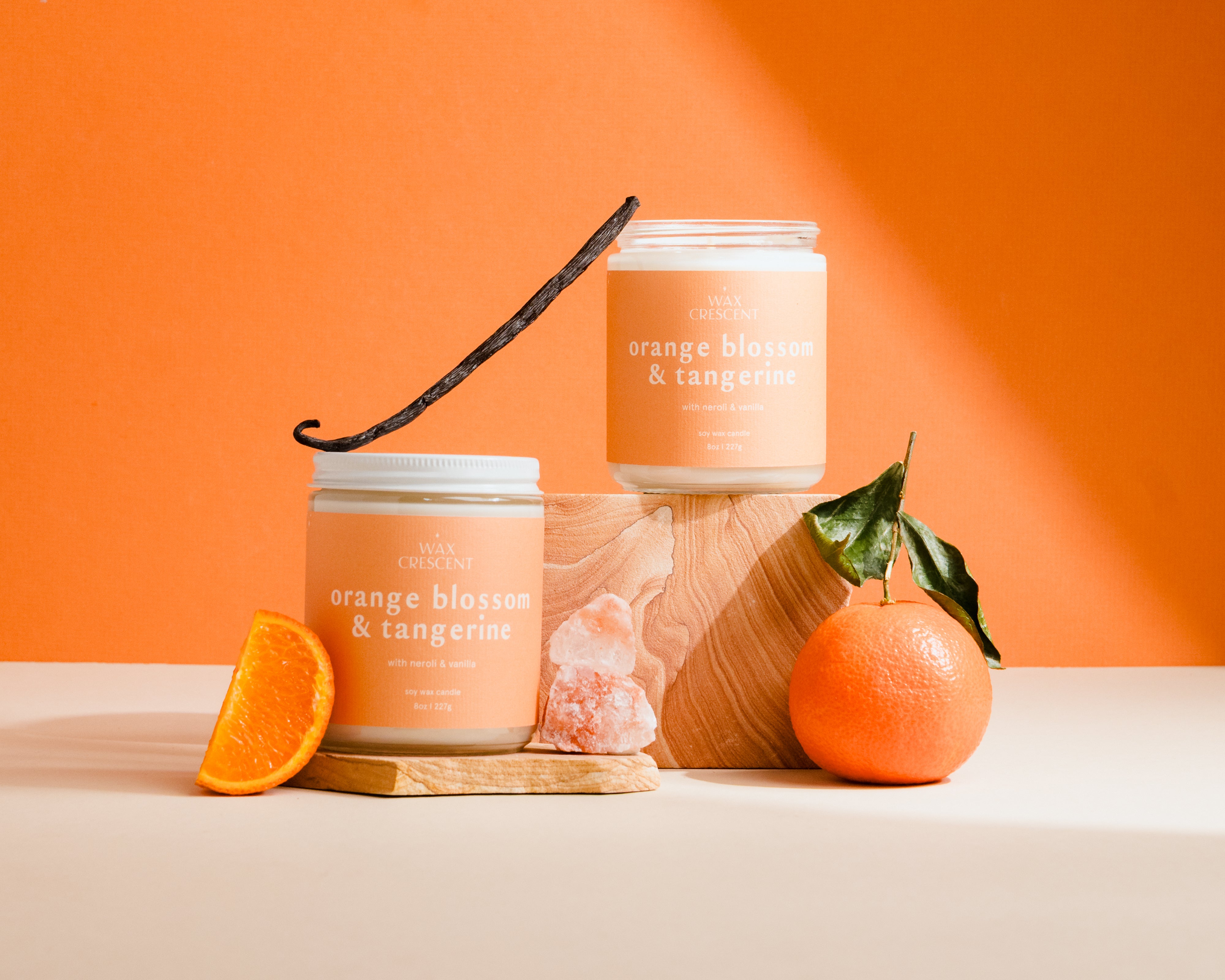 Two candles labeled 'Orange Blossom & Tangerine' with an orange, vanilla bean, and leaf on a peach background.