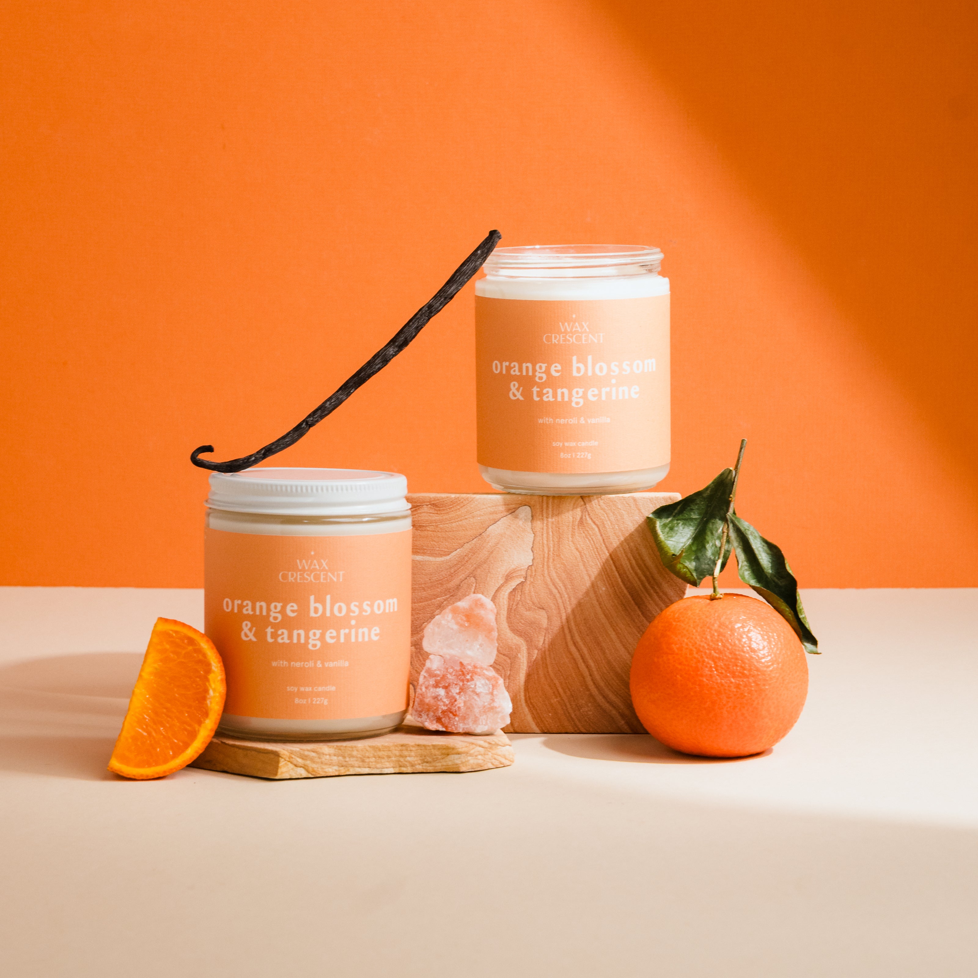 Two candles labeled 'Orange Blossom & Tangerine' with an orange, a vanilla bean, and a crystal on an orange background.