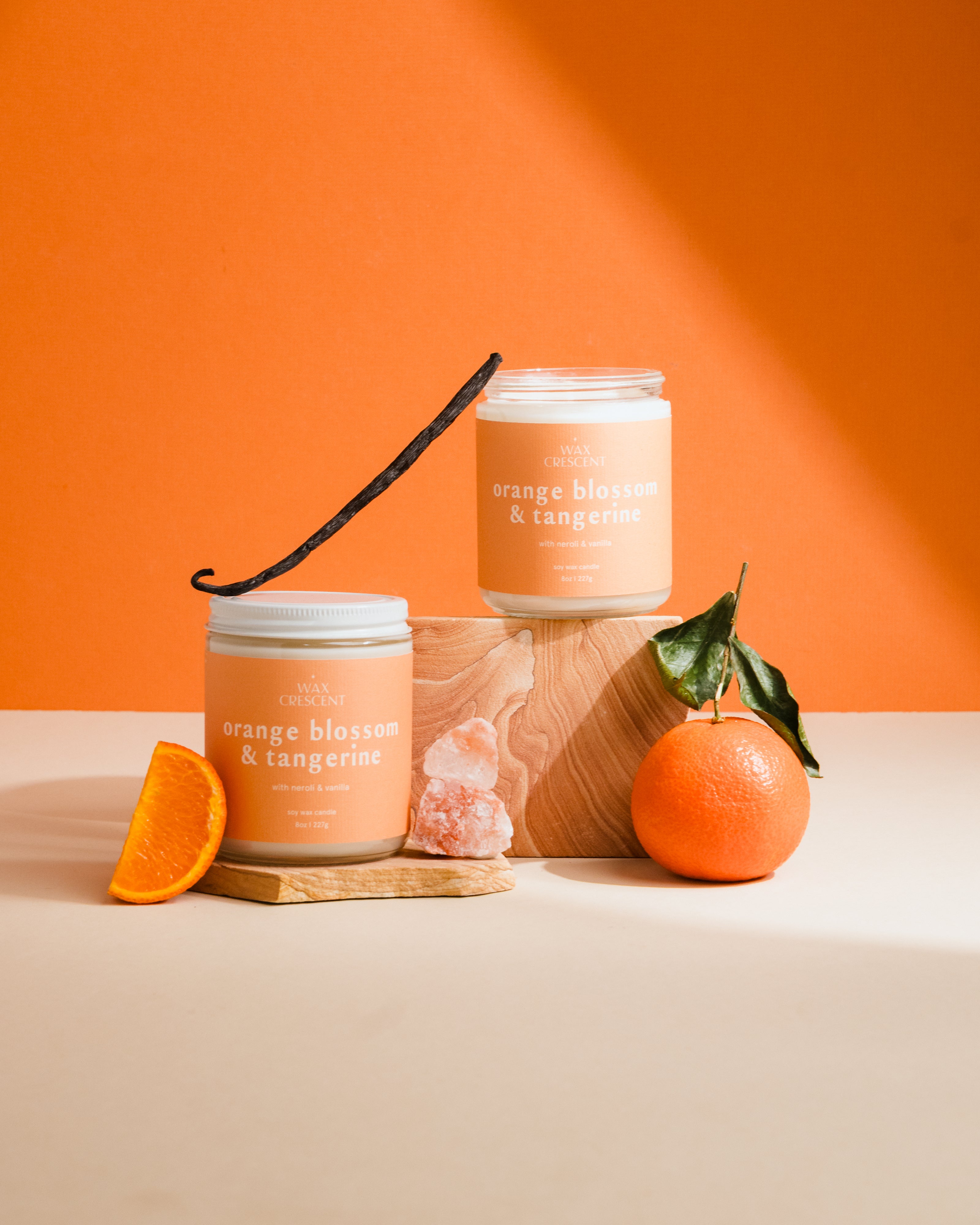 Two candles labeled 'Orange Blossom & Tangerine' with an orange, a vanilla bean, and a crystal on an orange background.