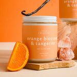Candle jar labeled 'Orange Blossom & Tangerine' with an orange slice and vanilla bean on a wooden board against an orange background.